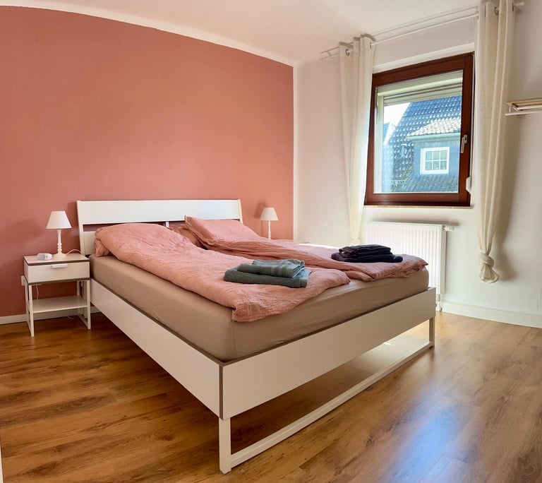 Modern bedroom with queen bed, coral accent wall, white bedding, pink nightstand lamp, wooden floor, and window with brown frame.